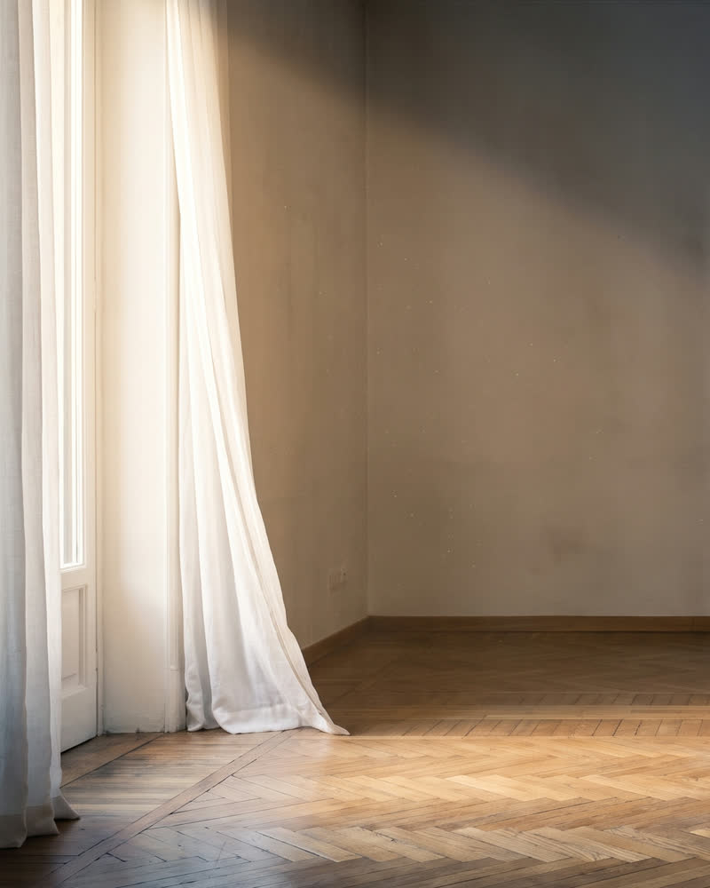 Tenda chiara e parquet, con la luce del mattino da una finestra alta.